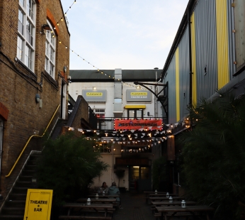 The exterior of Pleasance Islington. There is a courtyard with benches and a sign that reads 'theatre and bar'. There are stairs leading up to the entrance to the theatre and two large signs by the entrance that read Pleasance Theatre and Bar 
